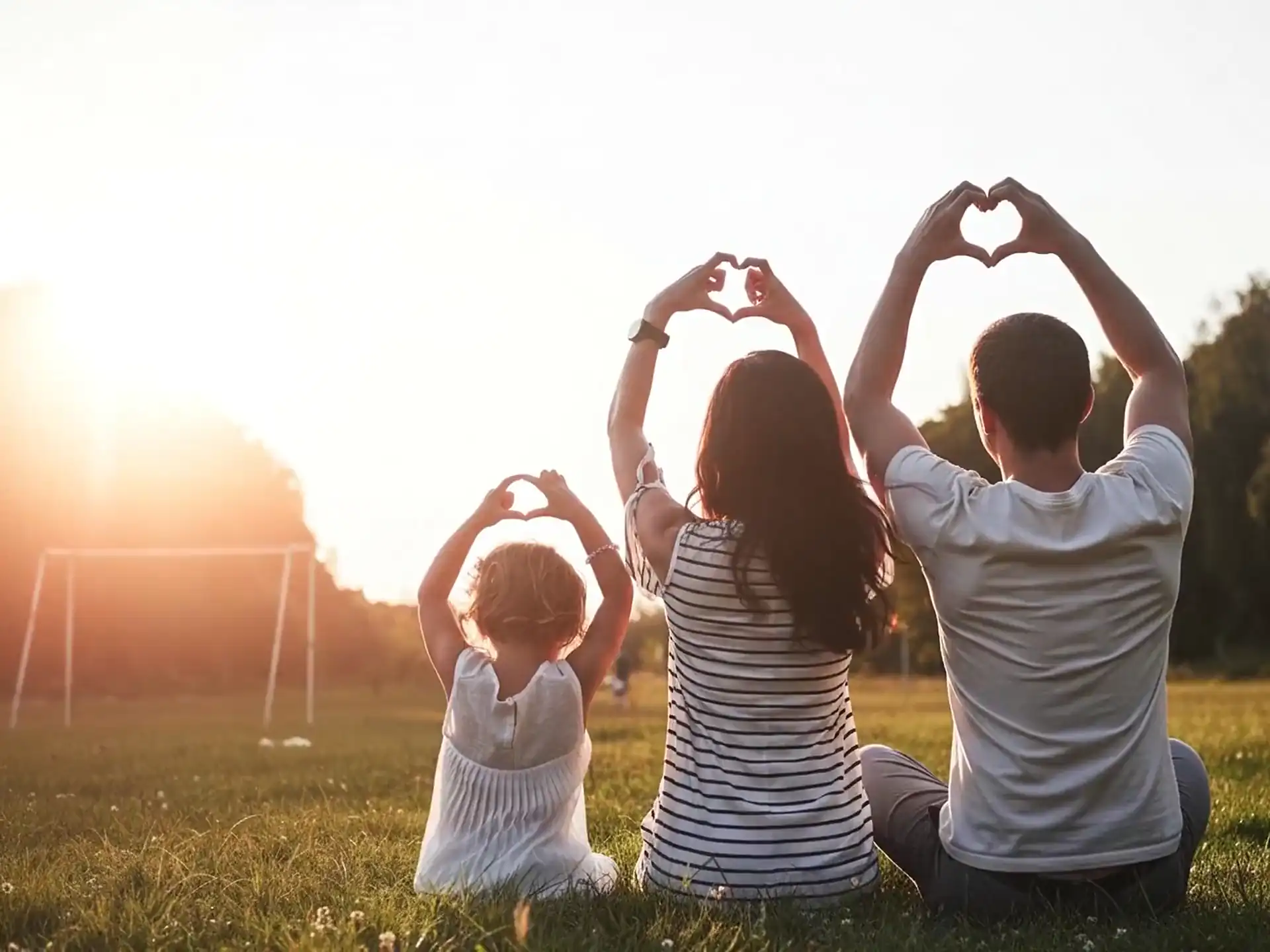 A family sitting on grass at sunset, making heart shapes with their hands
